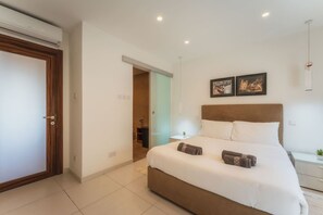 A bedroom with a large bed, a bedside table, a nightstand, a wall-mounted air conditioner, and a door. - Modern Apt in Marsaskala Near Seafront Beaches (Marsaskala)