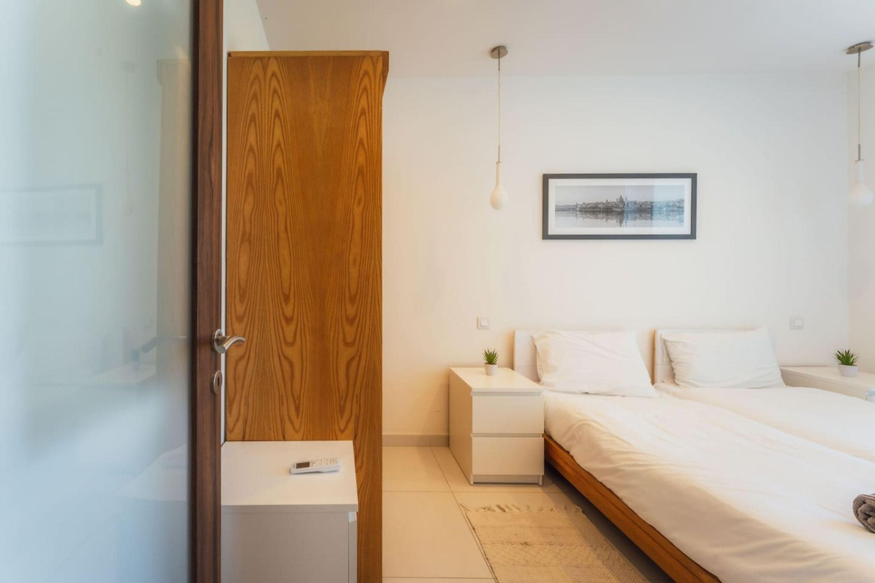 Quarto moderno com guarda-roupa de madeira, mesa de cabeceira com abajur, quadro emoldurado na parede e cama com roupa de cama branca.