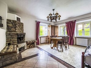 A room with a stone fireplace, a dining table with chairs, a chandelier, and large windows with curtains.