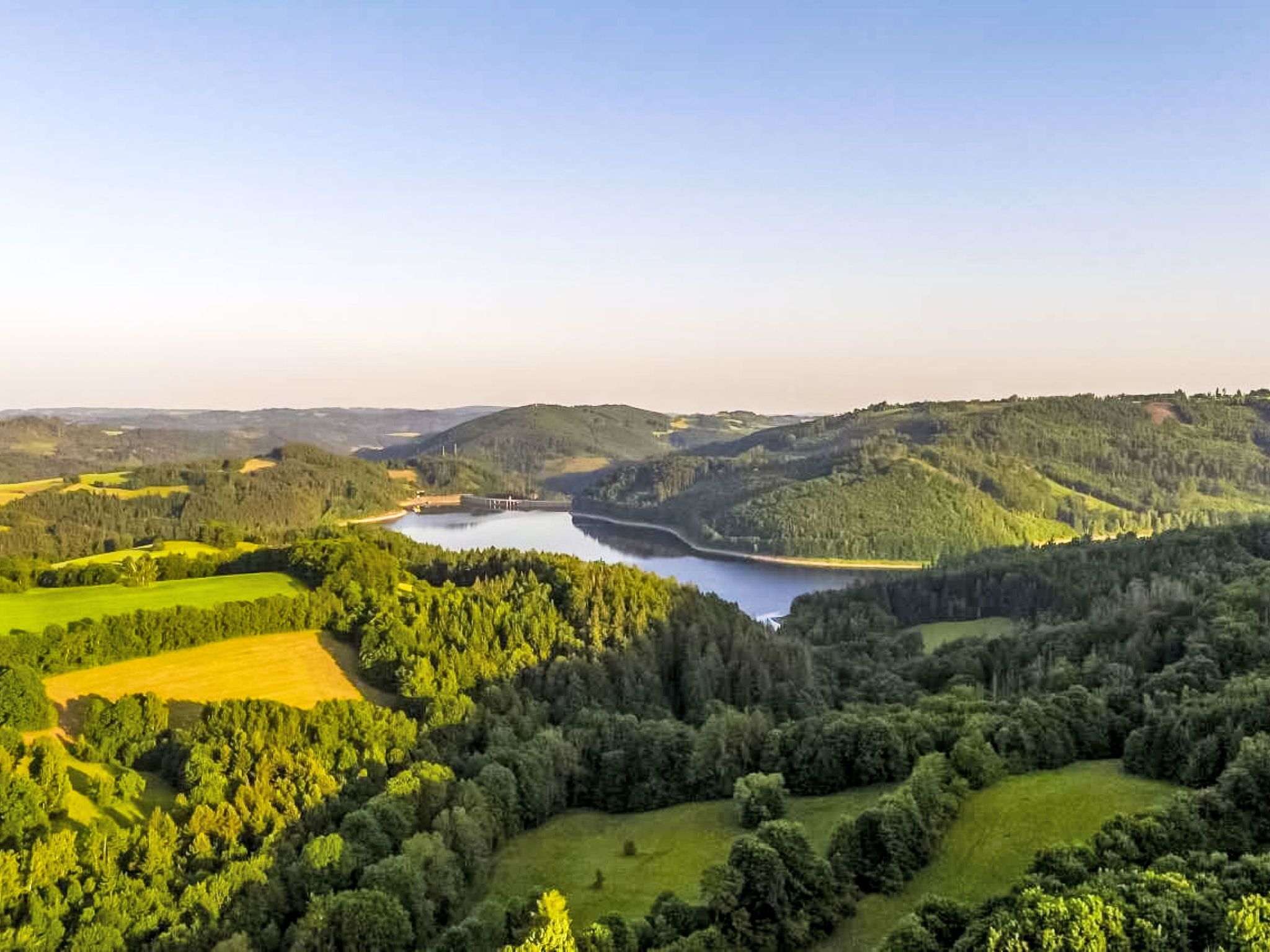 A panoramic view of a valley with a lake, surrounded by green fields and forested hills.