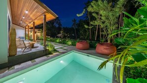 Un área de piscina moderna con terraza de madera, asientos y exuberante vegetación tropical.