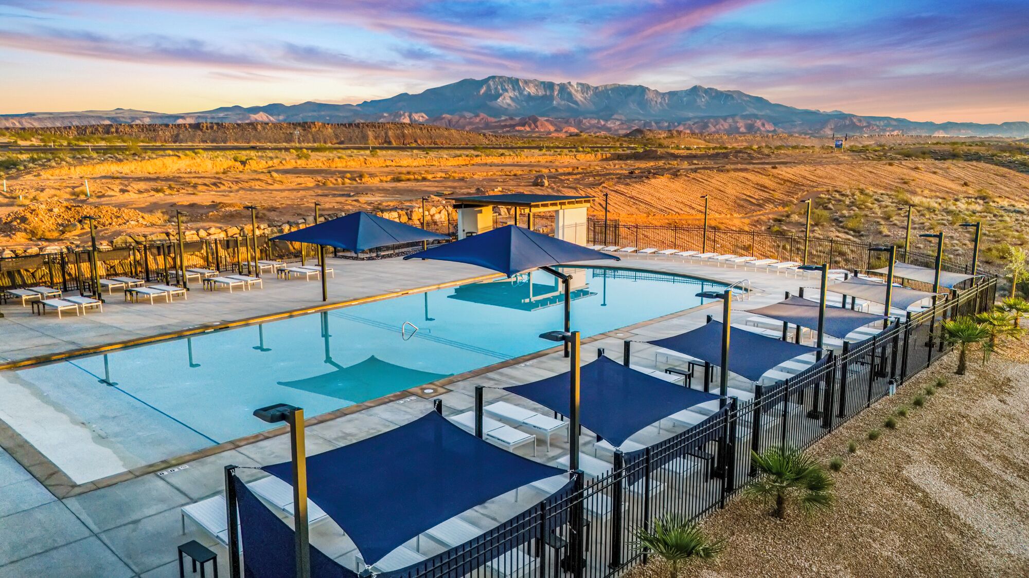 Zona de piscina con sillas de descanso y sombrillas, rodeada por una valla, con montañas al fondo.