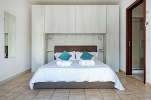Ein ordentlich bezogenes Bett mit weißer Bettwäsche und zwei türkisfarbenen Kissen, ein hölzernes Kopfteil und ein gefliester Boden.