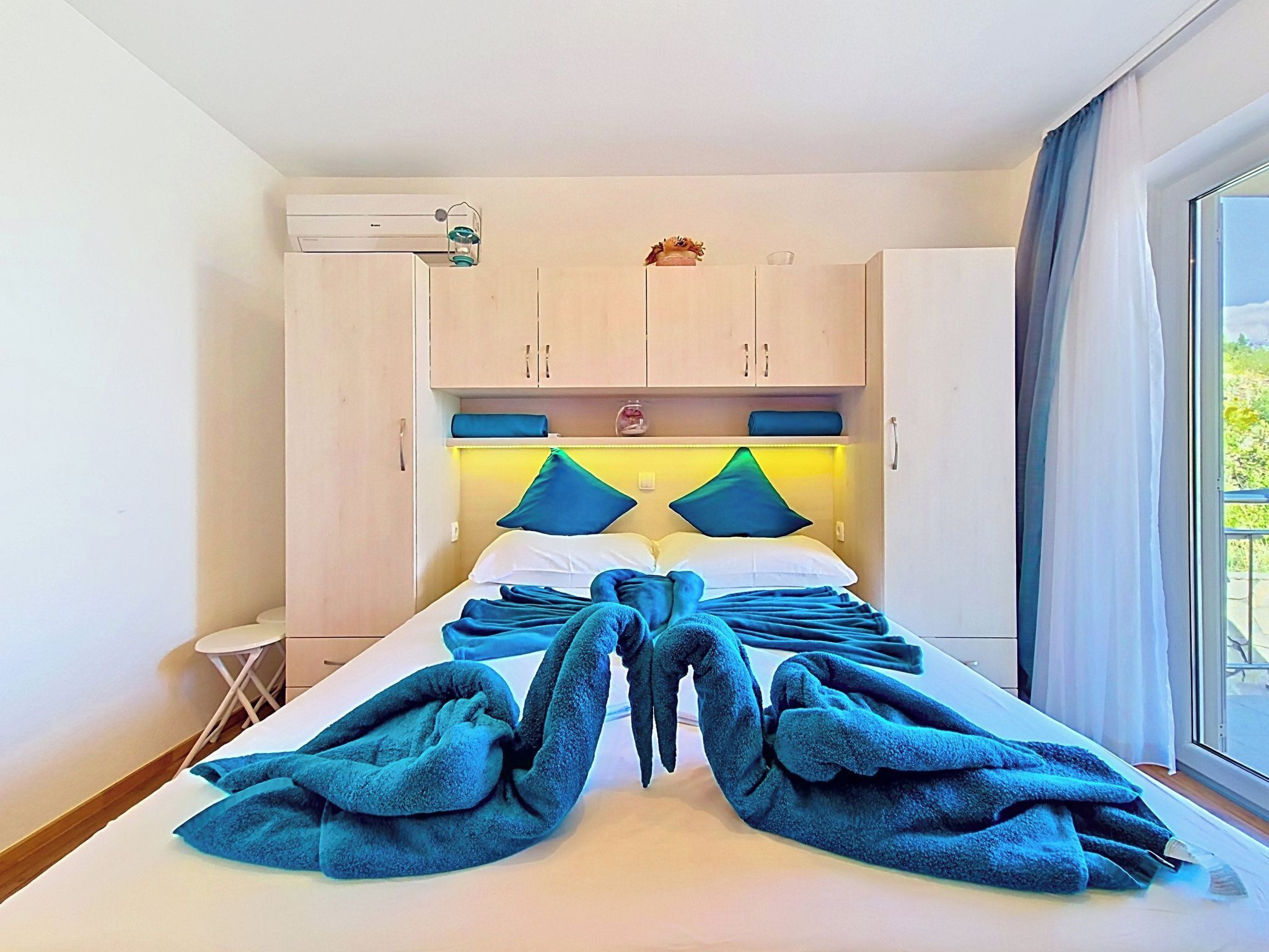 A hotel room with a bed, a small table, a window with curtains, and a ceiling-mounted air conditioner.