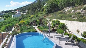Ein Schwimmbecken mit Liegestühlen, umgeben von einem Garten und einer Steinmauer, mit Häusern und Bergen im Hintergrund.