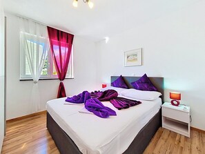 A neatly made bed with purple blankets, a bedside table with a lamp, and a window with curtains. - Apartment Palma by Interhome (Stanići)