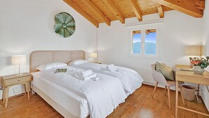 A bedroom with a wooden ceiling, a bed with white linens, a bedside table with a lamp, a chair, and a desk with a vase of flowers. - Agra Apartment (Lugano), Agra -Lugano, Switzerland (Agra -Lugano)