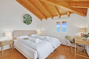 A bedroom with a wooden ceiling, a bed with white linens, a bedside table with a lamp, a chair, and a desk with a vase of flowers. - Agra Apartment (Lugano), Agra -Lugano, Switzerland (Agra -Lugano)