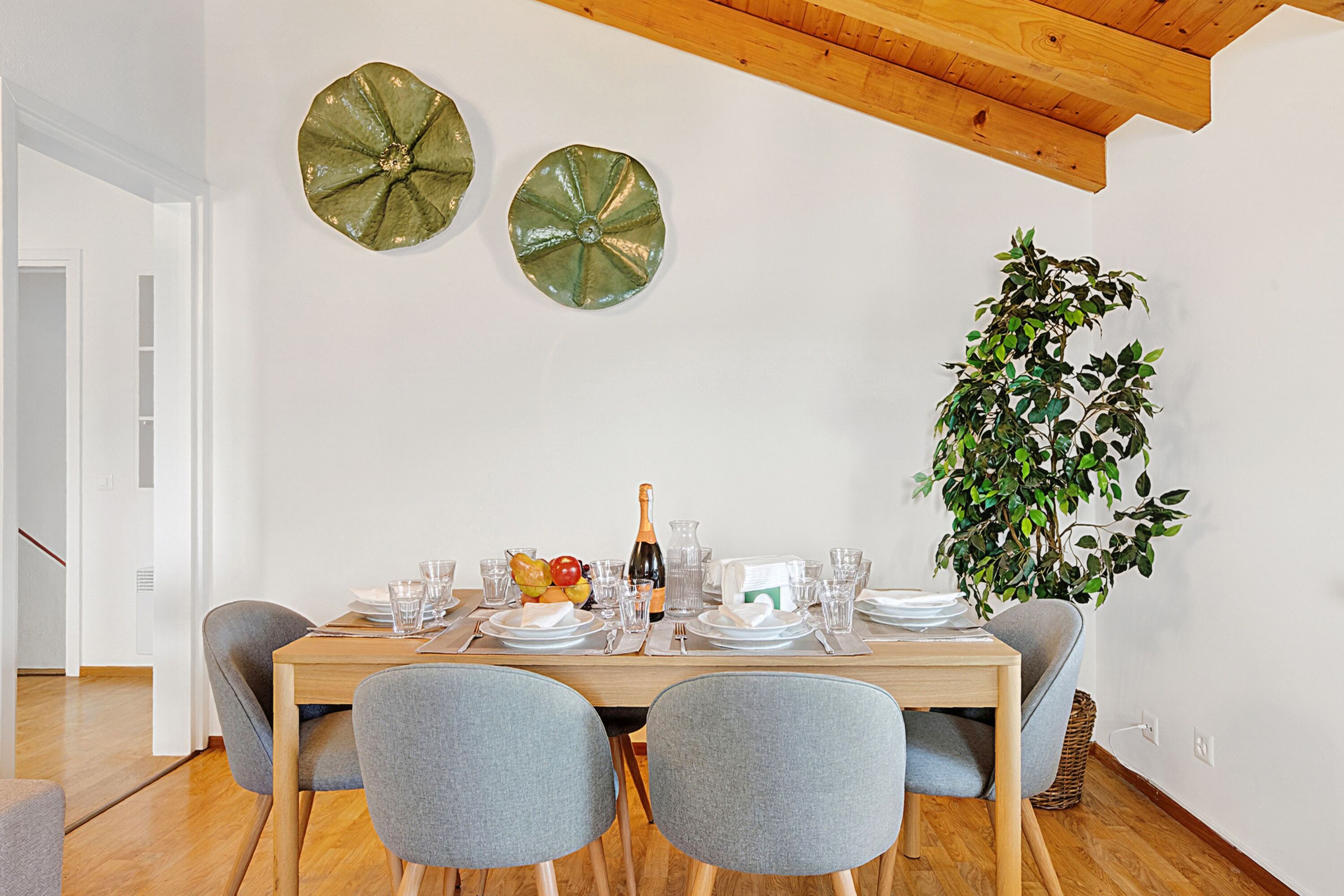 A dining area with a wooden table set for two, a potted plant, and decorative wall art.