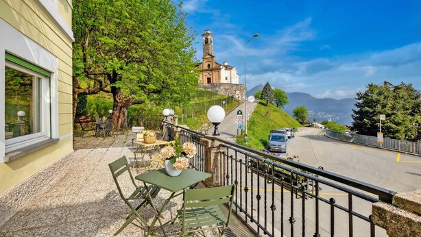 A balcony with outdoor seating, a view of a church, and a road with cars. - Agra 8 (Lugano), double, PrivateBathroom, Agra -Lugano, Switzerland (Agra -Lugano)