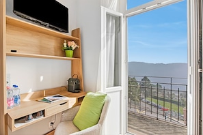 Une chambre dotĂ©e dâun bureau, dâune tĂ©lĂ©vision, dâune fenĂȘtre donnant sur un parking et dâun balcon avec une rambarde.