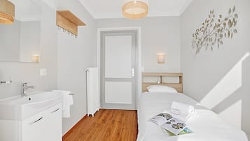 Ein Hotelzimmer mit Holzboden, weißen Wänden, einem großen Spiegel, einem weißen Waschbecken, einem Bett mit weißer Bettwäsche und einer Wandleuchte.