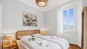 Hotellihuone, jossa on kaksi sänkyä, ikkuna, josta on näkymä, taideteoksilla koristeltu seinä sekä yöpöytä, jossa on lamppu.