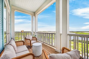 A covered porch with wicker furniture, a round white table, and a view of a grassy area and a distant building. - Multilevel beach house with water view, balconies, WIFI, kitchen and shared pool (Port Aransas)