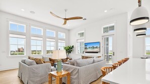 Amplia sala de estar con un sofá grande, una mesa de centro de madera, televisor y una zona de cocina con una isla blanca y taburetes de bar.