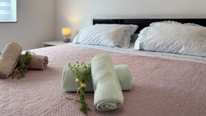 Ein ordentlich bezogenes Bett mit aufgerollten Handtüchern, einer kleinen Pflanze, einer Nachttischlampe und einem gerahmten Bild an der Wand.
