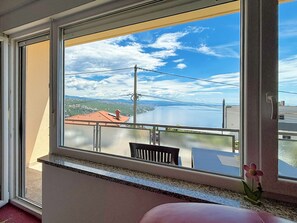 Ein Zimmer mit großem Fenster mit Blick auf einen See und Gebäude, ein Balkon mit einem Stuhl und einer Blumenvase.