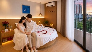 Una pareja sentada en una cama con un adorno con forma de corazón, una mesita de noche con un florero y vista a la ciudad.