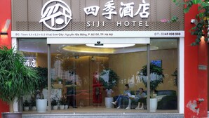 Entrada de hotel con fachada de vidrio, plantas en macetas y un área de recepción con asientos.