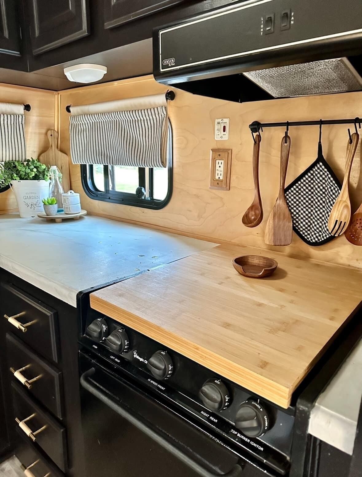 Una cocina compacta con encimera de madera, una estufa con varias hornillas, una ventana con cortinas a rayas y utensilios de madera colgando sobre la encimera.