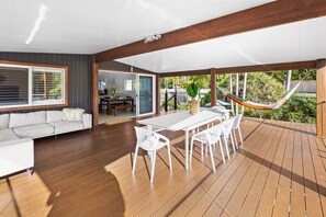Un espacio al aire libre techado con terraza de madera, mobiliario blanco y una hamaca.