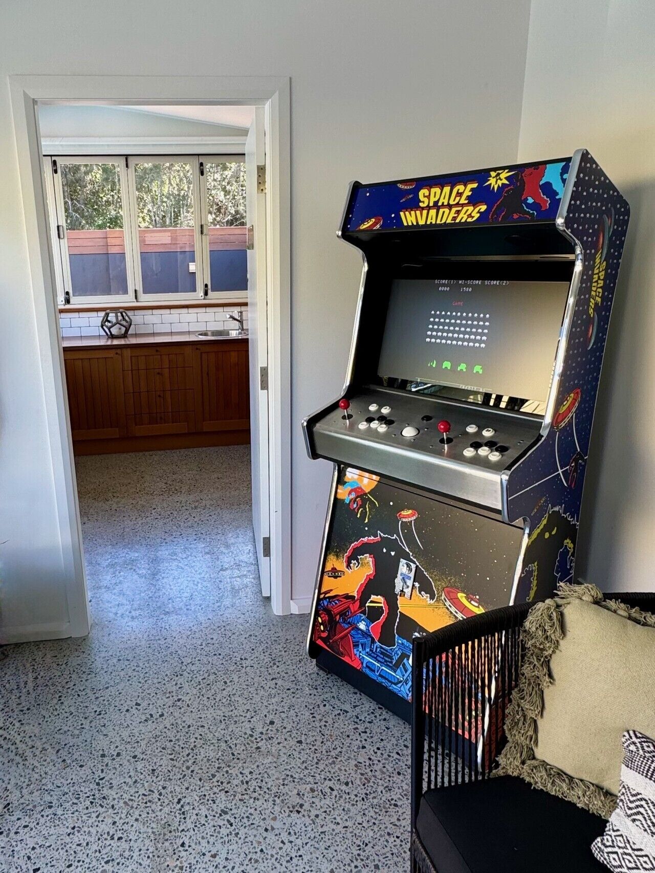 Una habitación con una máquina de juegos de salón con temática espacial, una ventana con persianas y una puerta que da a otra habitación.