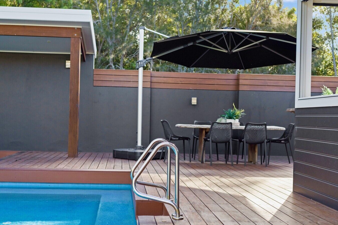 A poolside area with a table and chairs, a pool, and a pergola with an umbrella.