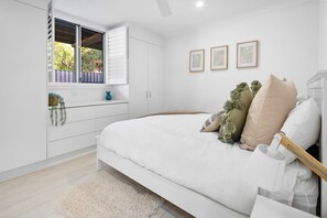 A bedroom with a bed, a window with blinds, a bedside table, and a wall-mounted shelf with plants.