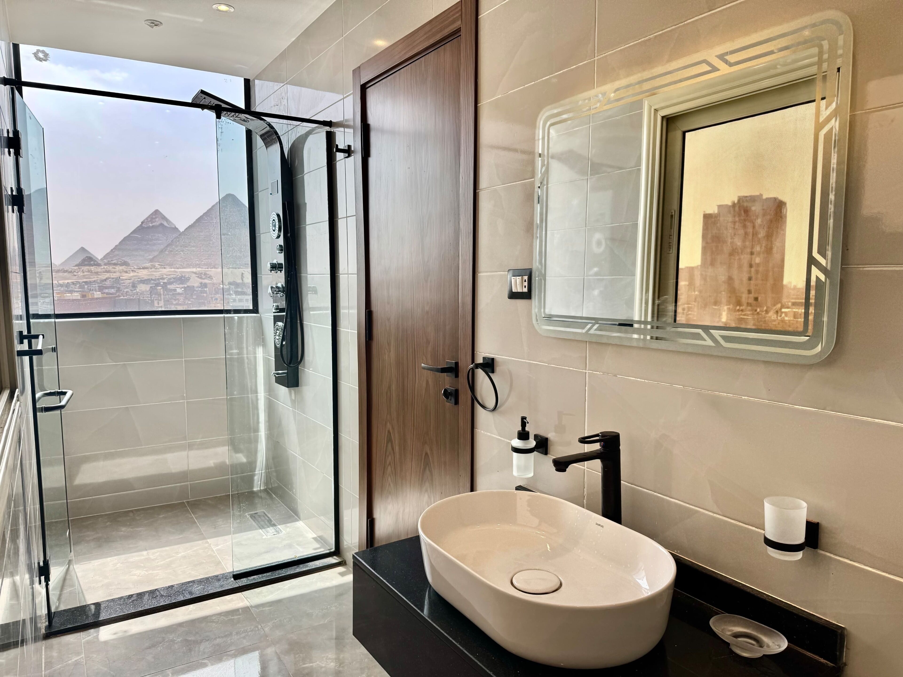A modern bathroom with a glass shower, a sink with a basin, a mirror with a cityscape reflection, and a view of pyramids through the door.. Cozy 1-private bedroom in heart of Giza pyramids 5 min walking
