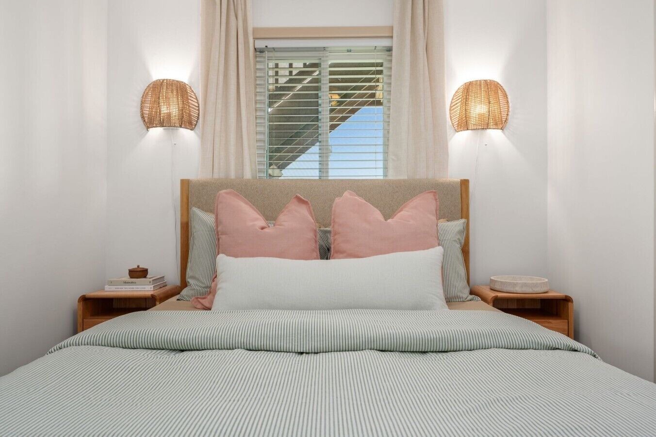 A bed with striped bedding, a headboard with decorative pillows, bedside tables with lamps, and a window with blinds.