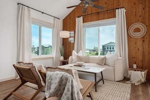 A room with a wooden ceiling, a ceiling fan, a sofa, two chairs, a table, and a view of buildings outside.