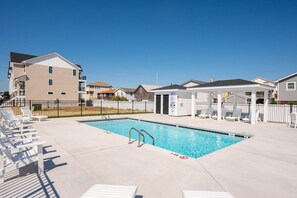 A swimming pool with lounge chairs, a covered seating area, and a building with multiple floors. - New Beachside Home! Pool & Pet Friendly! (Kill Devil Hills)