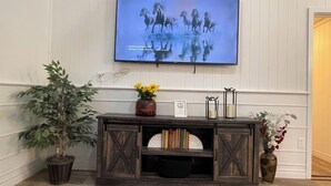 A television mounted on a wall displaying a horse race, a wooden console with decorative items, and a potted plant. - Spacious Country Getaway (Guilford)