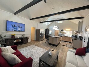 A spacious living area with a large TV, wooden flooring, a red sofa, a coffee table, and a kitchen in the background.