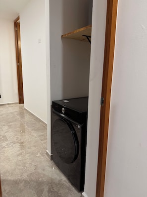 A laundry room with a washing machine, a wooden shelf, and a tiled floor.