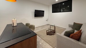 A modern living room with a flat-screen TV, a kitchen area with a stove, a sofa with cushions, and a hanging pendant light. - Luxe Apt. in Montebello Pool/Game Room/ Workspace (Mérida)