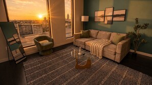 Ein Wohnzimmer mit Sofa, Sessel und Couchtisch, großen Fenstern mit Blick auf die Stadt und Wandbildern.