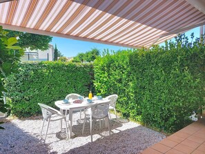 A covered outdoor dining area with a table and chairs, surrounded by a hedge and a building in the background. - Les Jardins de l'Océan by Interhome (Vaux Sur Mer)