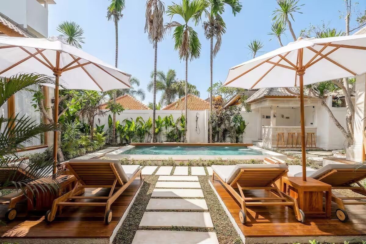 Un espace piscine agrémenté de chaises longues et de parasols, entouré de palmiers et surplombé d’un ciel dégagé.
