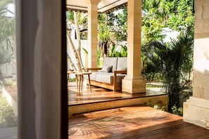 Una terraza de madera con un sofá y una mesa, rodeada de follaje y columnas.