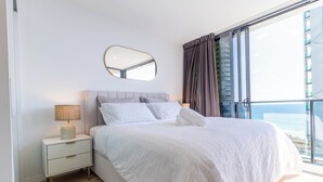Ein Schlafzimmer mit Bett, Nachttisch, Lampe und einem großen Fenster mit Vorhängen.