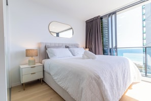 Ein Schlafzimmer mit Bett, Nachttisch, Lampe und einem großen Fenster mit Vorhängen.