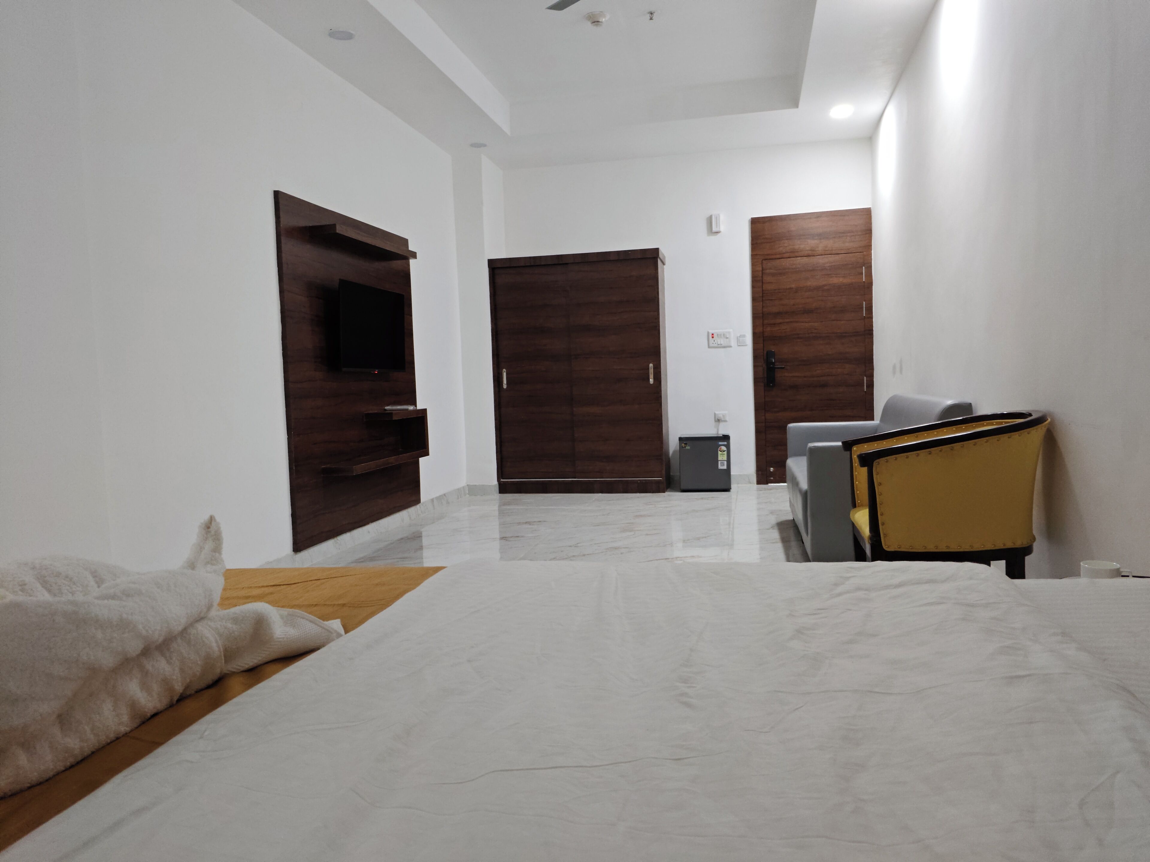 A hotel room with a bed, a television mounted on the wall, a wooden wardrobe, and a small table with a chair.