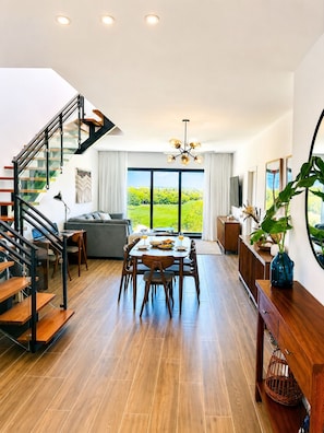 Ein modernes Wohnzimmer mit Essbereich, einer Treppe und großen Fenstern mit Blick auf eine grüne Landschaft.