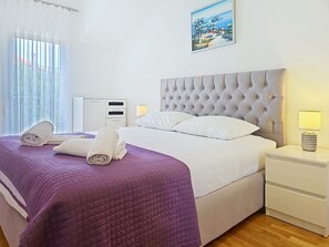 A neatly made bed with a purple quilt, white linens, and two rolled towels. There is a white bedside table with a lamp, a framed picture on the wall, and a window with sheer curtains. - Mary & Klara by Interhome (Trogir/Okrug Gornji)