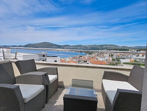 Eine Terrasse mit Korbmöbeln und einem Glastisch, mit Blick auf eine Küstenstadt und Hügel.