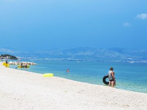 Ein Strand mit klarem Wasser, einer felsigen Küste und Bergen im Hintergrund.