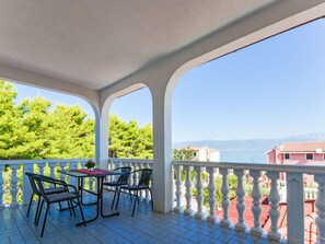 Ein überdachter Außenbereich mit Tisch und Stühlen, Blick auf das Meer und ein Gebäude im Hintergrund.