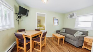 A room with a dining table, chairs, a sofa, a TV mounted on the wall, and a window. - 1 BR Suite 9 at Green Gables Resort (Osoyoos)