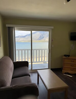 A room with a sofa, a table, a TV, and a view of a lake and mountains through a sliding door.